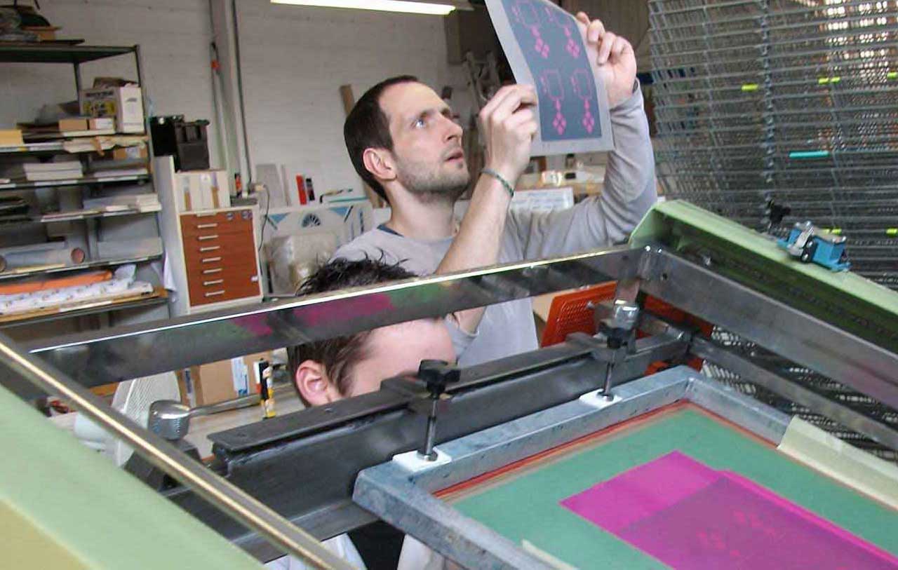 Tirage sur une machine de sérigraphie semi-automatique. Raphaël au tirage (margeage des feuilles de Lexan), et Vincent Benzi au contrôle et à la sortie des feuilles, avant lise sur claies pour séchage